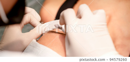 The hands of the cosmetologist are gluing white tape under the eye of the young caucasian woman during the eyelash extension procedure, closeup The hands of the cosmetologist are gluing white tape under the eye of the young caucasian woman during the eyelash extension procedure, closeup 94579260