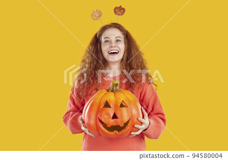 Portrait of cheerful young woman holding Halloween pumpkin with funny carved face on it. 94580004