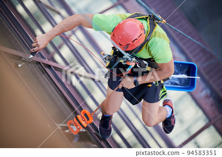 Industrial mountaineering worker washing glass windows of high-rise building, hanging on safety climbing rope. Man window cleaner in protective helmet cleaning skyscraper facade. Top view. 94583351