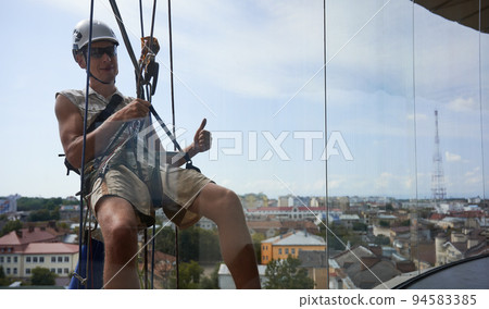 Industrial mountaineering worker in protective helmet showing approval gesture and smiling while hanging on rope behind window. Man using safety lifting equipment outside building. Industrial mountaineering worker in protective helmet showing approval gesture and smiling while hanging on rope behind window. Man using safety lifting equipment outside building. 94583385