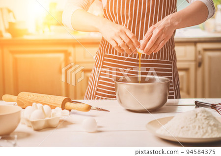 Young woman breaking egg over bowl with dough, close-up Young woman breaking egg over bowl with dough, close-up 94584375