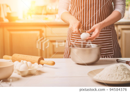 Young woman breaking egg over bowl with dough, close-up Young woman breaking egg over bowl with dough, close-up 94584376