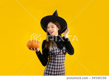 Halloween theme, young asian woman in black dress wearing witch hat holding pumpkin posing on yellow color background.. 94586198