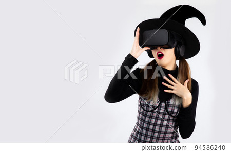 Halloween theme, young asian girl in black dress like witch costume wearing black witches hat and vr googles glasses watching and playing on white background.. Halloween theme, young asian girl in black dress like witch costume wearing black witches hat and vr googles glasses watching and playing on white background.. 94586240