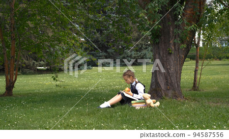 Little girl in school uniform reading a book while sitting on grass in a park. Literature, education and back to school concept Little girl in school uniform reading a book while sitting on grass in a park. Literature, education and back to school concept 94587556
