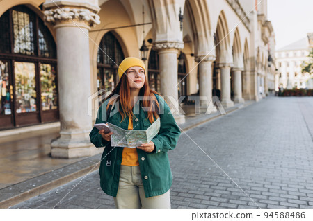 Attractive young female tourist is exploring new city. Redhead girl in hat holding a paper map and smartphone on Market Square in Krakow. Traveling Europe in autumn. Active lifestyle concept 94588486