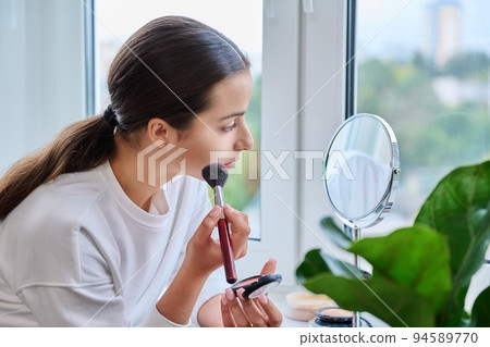 Young female with mirror doing morning daytime makeup, at home on windowsill 94589770
