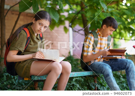 students a boy and a girl are sitting on a bench outside the school building, they are reading books students a boy and a girl are sitting on a bench outside the school building, they are reading books 94602073
