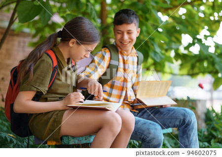 students a boy and a girl are sitting on a bench outside the school building, they are reading books and talking students a boy and a girl are sitting on a bench outside the school building, they are reading books and talking 94602075
