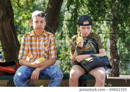 portrait of students in a city park, teenage schoolchildren a boy and a girl sitting on a bench, resting and eating apples 94602305