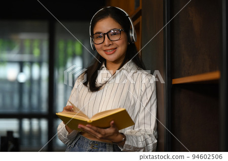 Positive female student reading a book between library bookshelves. Education and people concept 94602506