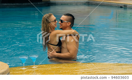 The loving couple hugs and kisses, drinking blue cocktail alcohol liquor in swimming pool at hotel outdoor. Portrait of caucasian man and woman. 94603603