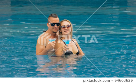 The loving couple hugs and kisses, drinking blue cocktail alcohol liquor in swimming pool at hotel outdoor. Portrait of caucasian man and woman. 94603629