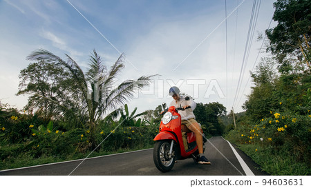 Man on red motorbike in white clothes drive on forest road trail trip. One men caucasian tourist go on scooter, nearby tropical palm tree. 94603631