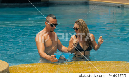 The loving couple hugs and kisses, drinking blue cocktail alcohol liquor in swimming pool at hotel outdoor. Portrait of caucasian man and woman. The loving couple hugs and kisses, drinking blue cocktail alcohol liquor in swimming pool at hotel outdoor. Portrait of caucasian man and woman. 94603755