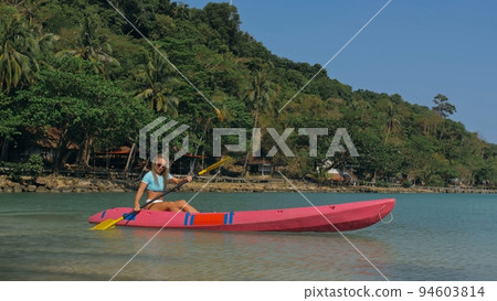 Young blonde woman in blue swimsuit rows pink plastic canoe along azure sea bay past island with palms under blue sky at resort. 94603814