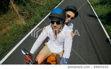 Two caucasian tourist woman man drive on red scooter. Make it selfie. Love couple on motorbike in white clothes to go on forest road trail trip. 94603830