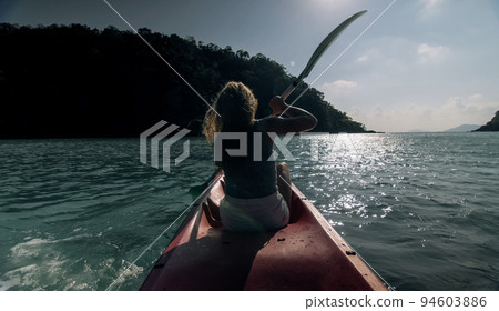 Silhouette of woman tourist raising hands while sailing in canoe along sea bay water to distant hilly island backside view. 94603886