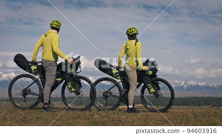 The man and woman travel on mixed terrain cycle touring with bikepacking. The two people journey with bicycle bags. Mountain snow capped. The man and woman travel on mixed terrain cycle touring with bikepacking. The two people journey with bicycle bags. Mountain snow capped. 94603916