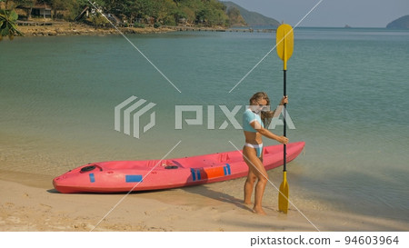 Pretty young sportswoman with sunglasses and swimsuit holds paddle posing near pink plastic canoe on sand beach at resort. 94603964