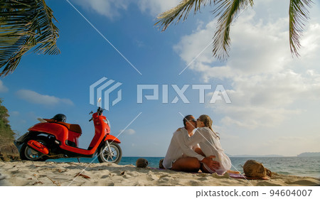 Scooter road trip. Lovely couple on red motorbike in white clothes on sand beach. People walking near the tropical palm trees, sea. Motorcycle rent. Scooter road trip. Lovely couple on red motorbike in white clothes on sand beach. People walking near the tropical palm trees, sea. Motorcycle rent. 94604007