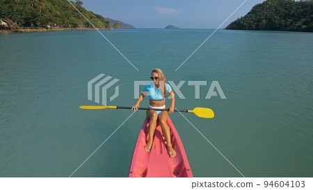 Pretty sportive blonde lady in stylish swimsuit sails on pink plastic canoe along azure ocean bay at exotic resort. Pretty sportive blonde lady in stylish swimsuit sails on pink plastic canoe along azure ocean bay at exotic resort. 94604103