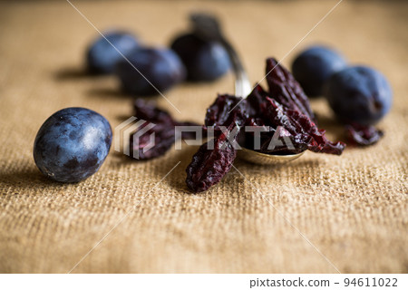 Dried prunes in a spoon on a burlap tablecloth against a background of fresh plums. 94611022