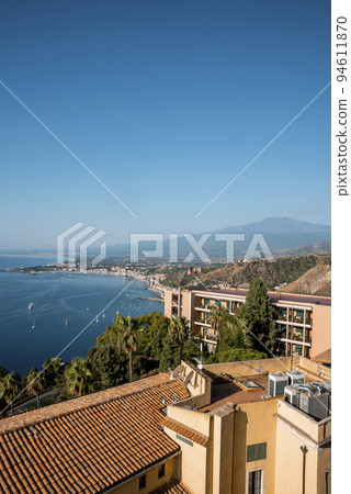 Idyllic view of volcanic Mount Etna and luxurious Elios hotel. Famous tourist attraction by seascape with blue sunny sky in background. Beautiful scenery of city during summer. 94611870