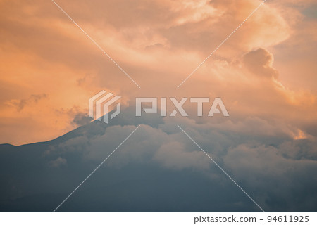 Scenic view of Mount Etna covered with smoke. Vibrant clouds wrapped around peak at twilight. Picturesque scenery of famous tourist attraction with orange sky in background during sunset. 94611925