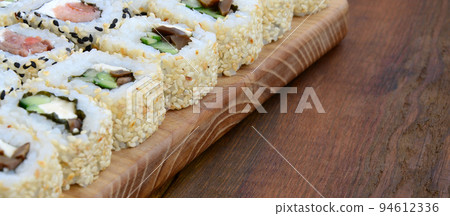 Close-up of a lot of sushi rolls with different fillings lie on a wooden surface. Macro shot of cooked classic Japanese food with a copy space Close-up of a lot of sushi rolls with different fillings lie on a wooden surface. Macro shot of cooked classic Japanese food with a copy space 94612336