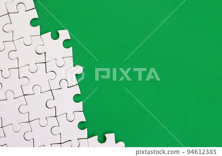 Fragment of a folded white jigsaw puzzle on the background of a green plastic surface. Texture photo with copy space for text Fragment of a folded white jigsaw puzzle on the background of a green plastic surface. Texture photo with copy space for text 94612385