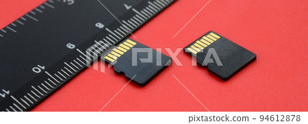 Two small micro SD memory cards lie on a red background next to a black ruler. A small and compact data and information store Two small micro SD memory cards lie on a red background next to a black ruler. A small and compact data and information store 94612878