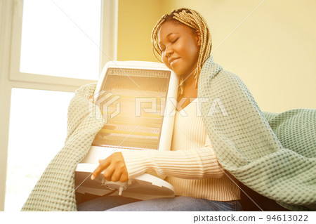 African american woman in blue blanket warming near electric heater at home African american woman in blue blanket warming near electric heater at home 94613022