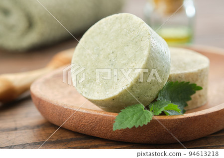 Nettle solid shampoo pieces or homemade natural organic soap bars on wooden soap dish, fresh green nettle leaves. A bath towel, essential oil bottle and comb on background. Selective focus. 94613218