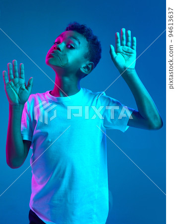 Close-up of little boy leaning against transparent glass by cheek isolated on blue background in neon light. Funny face. Concept of emotions Close-up of little boy leaning against transparent glass by cheek isolated on blue background in neon light. Funny face. Concept of emotions 94613637