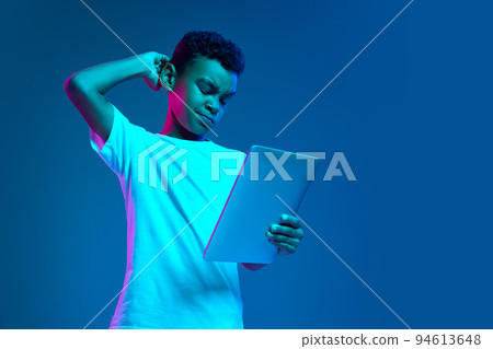 Serious little boy, kid in white t-shirt looking at tablet screen isolated over blue background in neon light. Remote education, childhood, modern lifestyle 94613648