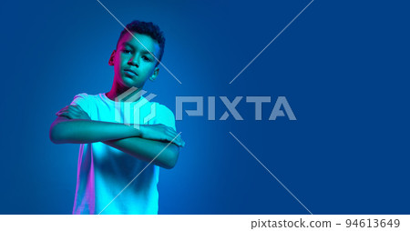 Studio shot of emotional little boy, kid standing with crossed hands isolated over blue background in neon light. Education, childhood, modern lifestyle 94613649