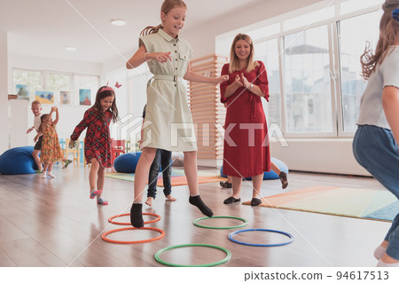 Small nursery school children with female teacher on floor indoors in classroom, doing exercise. Jumping over hula hoop circles track on the floor. Small nursery school children with female teacher on floor indoors in classroom, doing exercise. Jumping over hula hoop circles track on the floor. 94617513