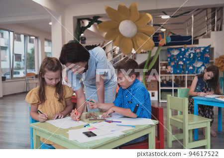 Creative kids during an art class in a daycare center or elementary school classroom drawing with female teacher.  94617522