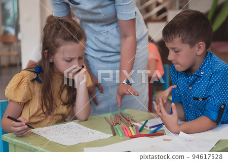 Creative kids during an art class in a daycare center or elementary school classroom drawing with female teacher.  94617528
