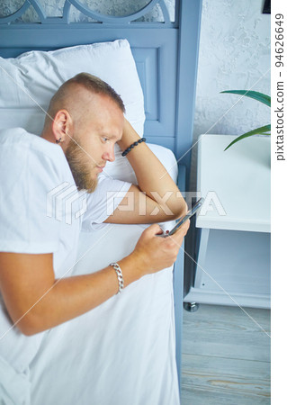 Bearded man using smartphone in morning in cozy while lying on bed Bearded man using smartphone in morning in cozy while lying on bed 94626649