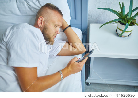 Bearded man using smartphone in morning in cozy while lying on bed Bearded man using smartphone in morning in cozy while lying on bed 94626650
