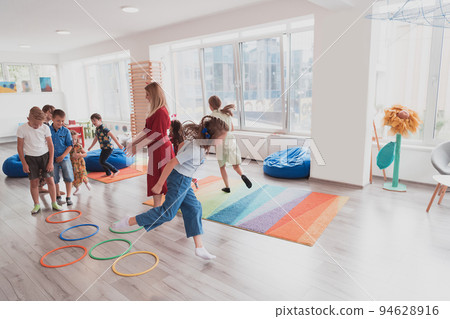 Small nursery school children with female teacher on floor indoors in classroom, doing exercise. Jumping over hula hoop circles track on the floor. Small nursery school children with female teacher on floor indoors in classroom, doing exercise. Jumping over hula hoop circles track on the floor. 94628916