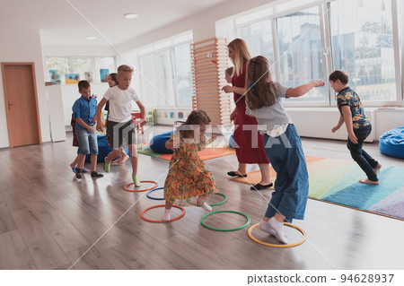 Small nursery school children with female teacher on floor indoors in classroom, doing exercise. Jumping over hula hoop circles track on the floor. 94628937