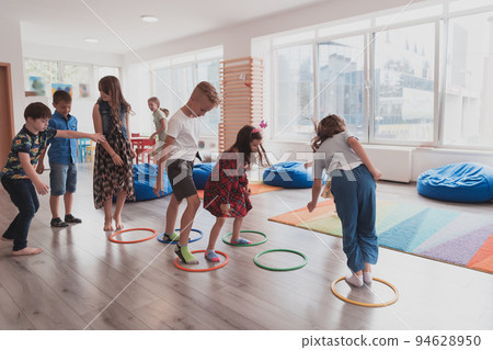 Small nursery school children with female teacher on floor indoors in classroom, doing exercise. Jumping over hula hoop circles track on the floor. 94628950