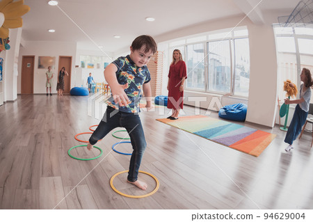 Small nursery school children with female teacher on floor indoors in classroom, doing exercise. Jumping over hula hoop circles track on the floor. Small nursery school children with female teacher on floor indoors in classroom, doing exercise. Jumping over hula hoop circles track on the floor. 94629004