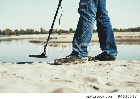 Man with metal detector walks along the sandy river bank. Search for treasures and metal for recycling Man with metal detector walks along the sandy river bank. Search for treasures and metal for recycling 94630588