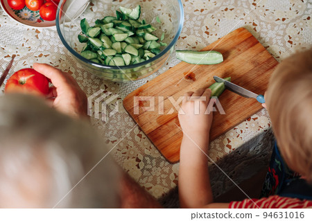 Young child cuts a cucumber carefully with a knife. Child learns to cut a cucumber on a cutting board. Young child cuts a cucumber carefully with a knife. Child learns to cut a cucumber on a cutting board. 94631016