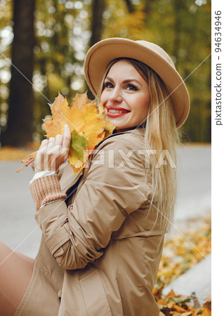 Woman posing for a photo in the autumn park. Caucasian girl has red lips. Woman wearing beige coat and a hat. 94634946
