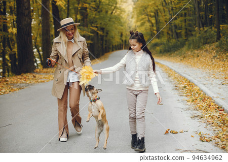 Woman, little girl and dog walking in the autumn park. Brown french bulldog with female owners spend a day at the park playing and having fun. Woman wearing beige coat and a hat. 94634962